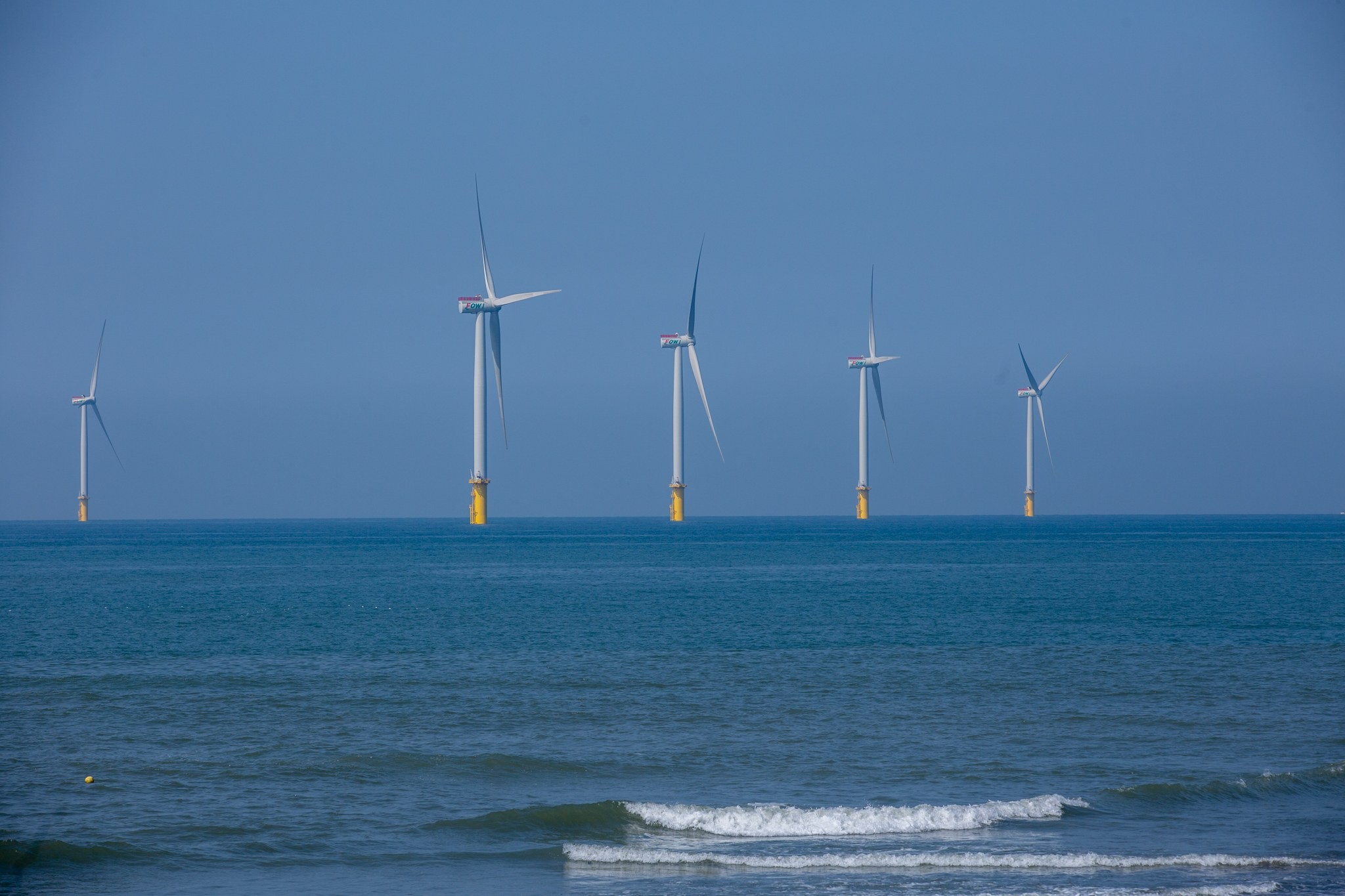 A row of Formosa 1 offshore wind turbines on a blue sea off Miaoli, with waves in the foreground