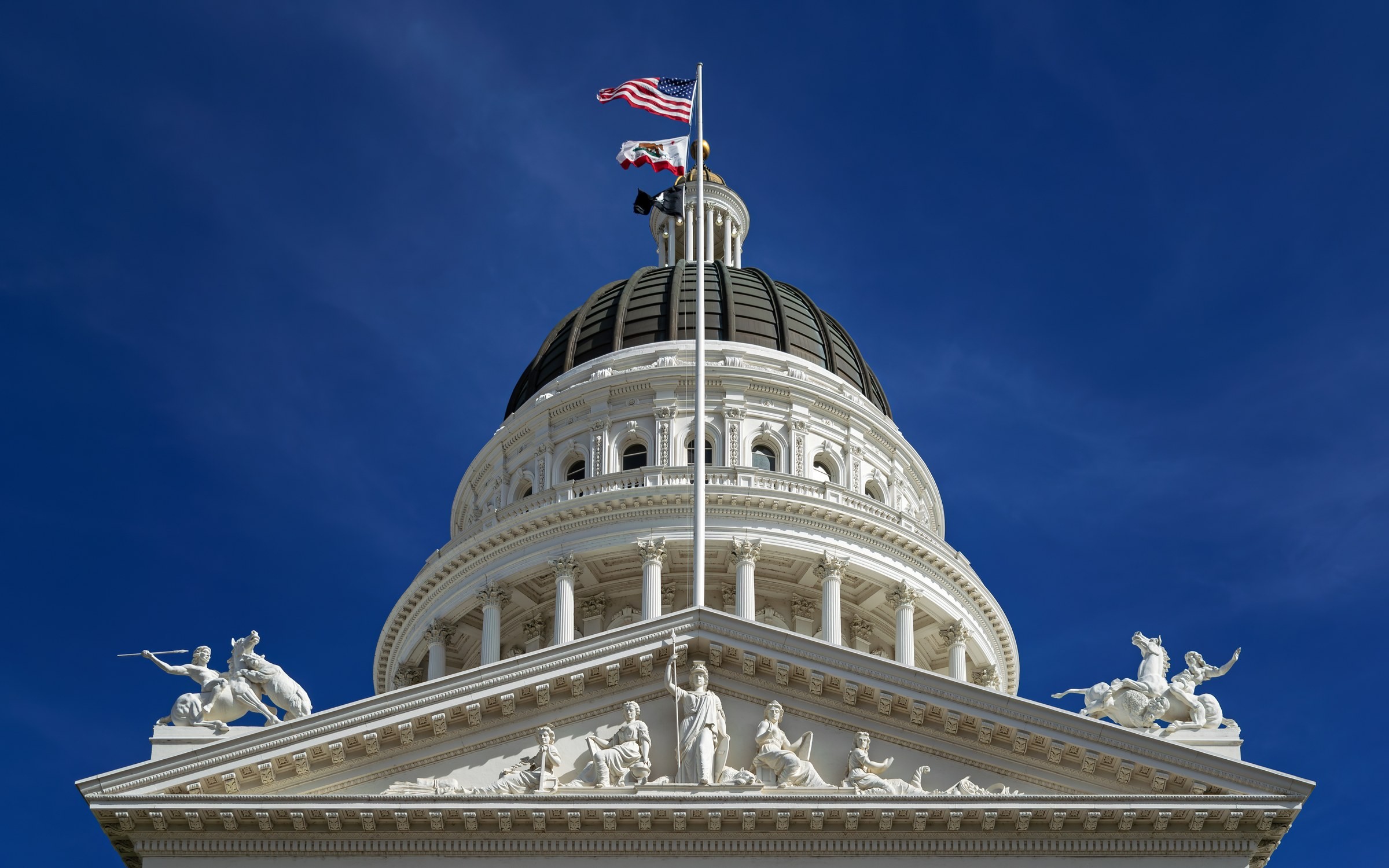 青空の下のカリフォルニア州議会議事堂のドームと破風。頂上に米国旗と州旗がはためく